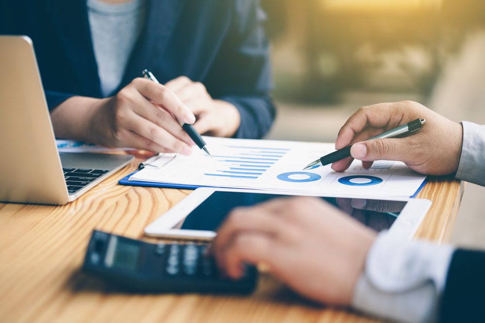 Financial management | Two people pointing at graphs on paper. One on a laptop and one using a calculator.